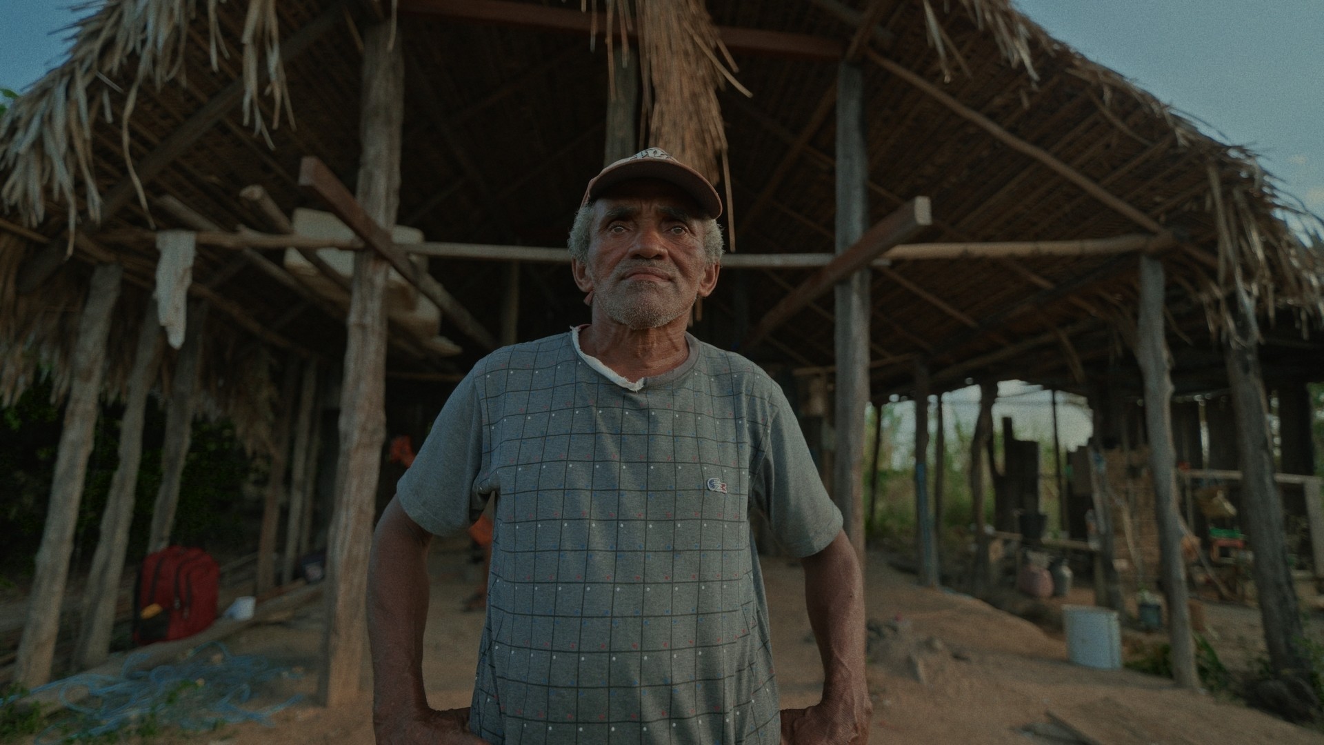 Pescador de meia-idade parado em frente ao seu barracão com telhado de palha e redes para dormir. Vestido com camisa velha cor cinza, calça velha cinza e boné velho cor vermelha com o brasão do Flamengo. É dia e o céu está com poucas nuvens Pescador de meia-idade parado em frente ao seu barracão com telhado de palha e redes para dormir. Vestido com camisa velha cor cinza, calça velha cinza e boné velho cor vermelha com o brasão do Flamengo. É dia e o céu está com poucas nuvens